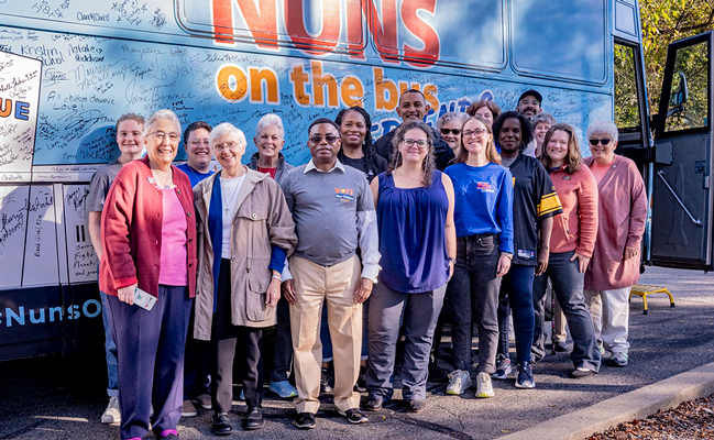 Sister Barbara Pfarr joins with others in a group picture in front of the Nuns on the Bus bus. The bus is covered in artwork and signatures from riders and participants at event stops.