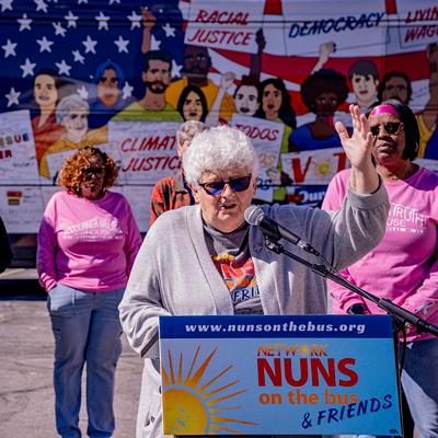 Sister Barbara Pfaff speaks during a Nuns on the Bus tour stop, with the bus parked behind her. 